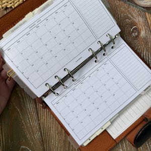 May include: Open planner with calendar pages for January and February 2026. The planner is brown leather with silver rings and dividers. The calendar pages are white with black text and dates.