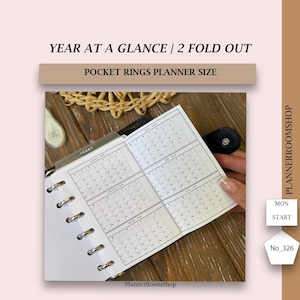以下が含まれることがあります： 年間カレンダー付きの白いポケットリングプランナーインサート。カレンダーは4つのセクションに分かれており、それぞれ3か月を表示しています。「YEAR AT A GLANCE / 2 FOLD OUT」の文字が上部にあります。プランナーは開いており、カレンダーページが表示されています。