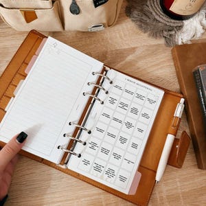 May include: A brown leather planner with a 5-minute decluttering checklist. The planner is open, revealing lined pages and a pen. A beige organizer with pens and a candle are in the background.