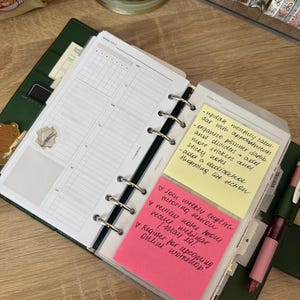 May include: A dark green ring-bound planner open to a weekly layout with white pages. A yellow sticky note lists tasks including updating a calendar and adding stickers. A pink sticky note below lists tutoring and workshop tasks in black ink.