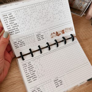 May include: Open planner with habit tracker pages. The pages are white with black grid lines and handwritten entries. A decorative tape with bear illustrations is across the pages. The planner is held open with black rings.