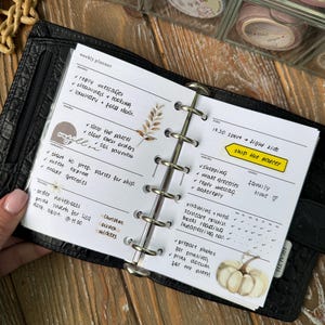 Pode incluir: Um planejador de couro preto aberto em uma página semanal. As páginas do planejador têm anotações manuscritas, caixas de seleção e adesivos decorativos. O texto inclui "ship the parcel" e "family time". Rolos de fita washi são visíveis no fundo.