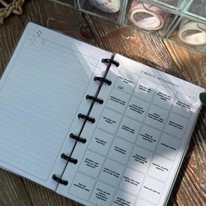 May include: An open planner with lined pages and a black binding. The right page features a "5 Minute Decluttering" checklist with various tasks. Decorative washi tape rolls are visible in the background, suggesting a craft or organization theme.