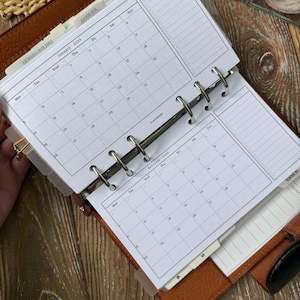 Pode incluir: Uma agenda de argolas em couro castanho com folhas brancas contendo calendários impressos de janeiro de 2026 e fevereiro de 2026. A agenda inclui divisórias com abas rotuladas com categorias como Journal, Skin Care e Online Orders.