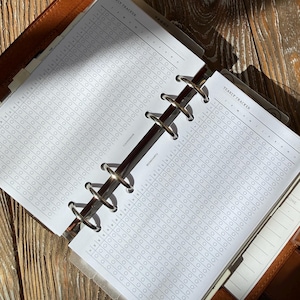 Puede incluir: Una agenda de anillas de cuero marrón con insertos de papel blanco que presentan una cuadrícula de seguimiento anual impresa. Las páginas incluyen columnas para meses y filas para días, con el texto Yearly Tracker y Tarot visible.
