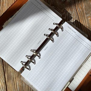 May include: Open brown leather planner with a yearly tracker insert. The insert is white with a grid of small circles for tracking. The planner is open, revealing the metal rings and the "Yearly Tracker" title at the top of the page. The background is a wooden surface.