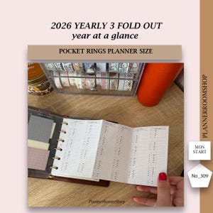 May include: A brown pocket rings planner with a 2026 yearly 3-fold-out calendar. The open planner displays a detailed schedule. The image also shows a clear container with decorative tapes and a tall orange container. Text on the image includes "PlannerRoomShop".