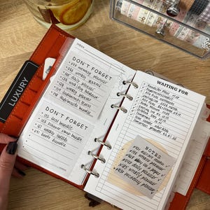 Könnte beinhalten: Ein rostfarbener Ringplaner aus Leder, aufgeschlagen auf Seiten mit handschriftlichen Listen unter den Überschriften DON'T FORGET, WAITING FOR und NOTES. Der Planer hat weißes liniertes Papier, silberne Metallringe und einen schwarzen Reiter mit LUXURY.