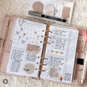 Op de afbeelding: Een open planner met een maandelijkse kalender, een to-do-lijst en een gewoontetracker. De planner is versierd met stickers en een bloemenontwerp. De tekst op de pagina omvat "Werk", "Factuur vervallen", "Kleding voor baby bestellen", "Boeken voor baby bestellen" en "Papierplanner verzenden".