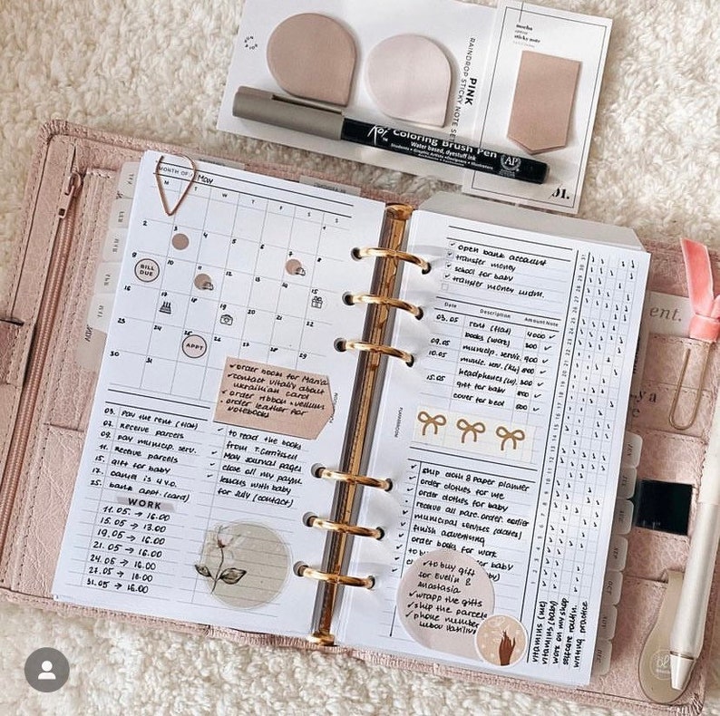 Op de afbeelding: Een open planner met een maandelijkse kalender, een to-do-lijst en een gewoontetracker. De planner is versierd met stickers en een bloemenontwerp. De tekst op de pagina omvat "Werk", "Factuur vervallen", "Kleding voor baby bestellen", "Boeken voor baby bestellen" en "Papierplanner verzenden".