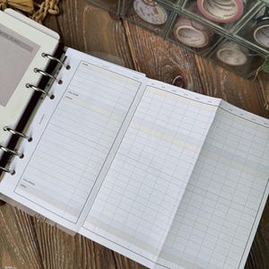 May include: Open planner with lined pages for financial tracking. The planner is open to a spread with columns for income, expenses, and savings. A clear container with decorative washi tape rolls is in the background.