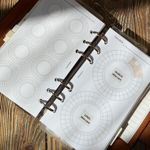 Könnte beinhalten: Ein Ringbuch-Planer mit weißen Seiten, die kreisförmige Tracker-Diagramme enthalten. Die Seiten zeigen gedruckte Beschriftungen wie TRACKER, TAROT, MARCH skin care und APRIL skin care. Einige Abschnitte der kreisförmigen Diagramme sind mit hellbrauner Tinte ausgefüllt.