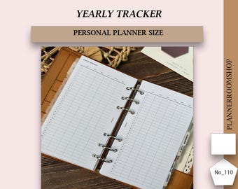 Può includere: Un planner annuale di dimensioni personali di colore bianco con una copertina in pelle marrone. Il planner è aperto su una pagina con una griglia per tenere traccia delle date e degli eventi. Il testo "YEARLY TRACKER" e "PERSONAL PLANNER SIZE" è stampato sulla copertina.