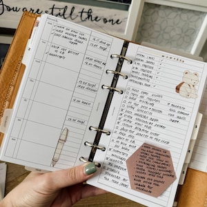 May include: A close-up of a weekly planner with a brown leather cover. The planner is open to the week of the month. The page has a list of tasks and appointments written in black ink. A sticky note with a list of tasks is attached to the page. The sticky note is pink with black ink. The text on the sticky note reads: "Make pictures of new products, add the items to the SCL (how to pack the new product), package covers, add inserts (printed),  excel another price list, create the parcel and sold out labels for products."