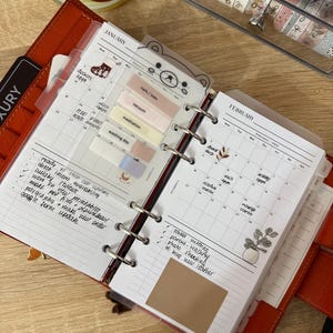 May include: A ring-bound planner open to January and February calendar pages. The pages feature handwritten tasks, colorful adhesive tabs, and decorative stickers including a bear, a plant, and text. The planner is held in a red leather cover.