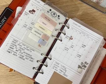 May include: A ring-bound planner open to January and February calendar pages. The pages feature handwritten tasks, colorful adhesive tabs, and decorative stickers including a bear, a plant, and text. The planner is held in a red leather cover.