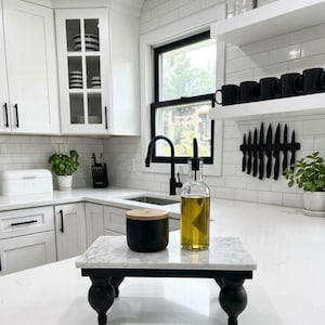 May include: A rectangular white marble riser with four black turned wooden feet. Sitting on the marble surface is a matte black cylindrical container with a light wood lid and a clear glass bottle filled with olive oil and a black pour spout.