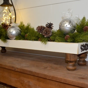 May include: White wooden tray with four turned wooden legs, decorated with greenery, pine cones, and silver Christmas baubles. The tray has a "Joy" sign on the side.