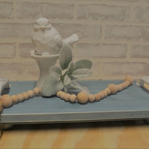 May include: A blue serving tray with white handles, decorated with a wooden bead garland and a white bird ornament. The tray also features faux greenery, creating a rustic, farmhouse aesthetic.