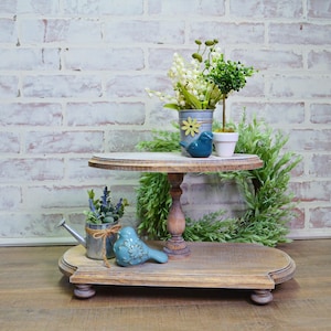 May include: Two-tiered wooden stand with a distressed white finish. The top tier features a small potted plant, a blue bird figurine, and a white pot. The bottom tier has a blue bird figurine, a small watering can, and a potted plant with lavender flowers.