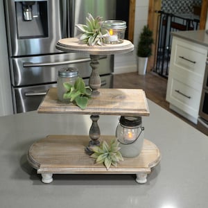 Puede incluir: Un soporte de servicio de madera de tres niveles con un acabado blanco desgastado. El soporte está decorado con vegetación y velas en frascos de vidrio.