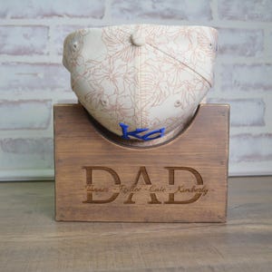 May include: A wooden hat display stand with the word "DAD" engraved on the front. The stand is holding a white baseball cap with a floral pattern and a blue "K" on the front. The names "Tanner, Rylie, Cate, Kimberly" are engraved below the word "DAD".