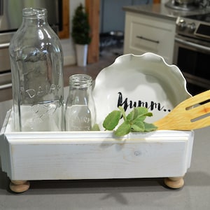 以下が含まれることがあります： 装飾的な縁取りと旋盤で仕上げられた脚が付いた白い木製の箱。箱の中には、透明なガラス製の牛乳瓶が2本、白い陶器のボウル（「Yumm...」の文字が書かれている）、木製のヘラが入っています。箱の中に緑色のハーブの枝があります。