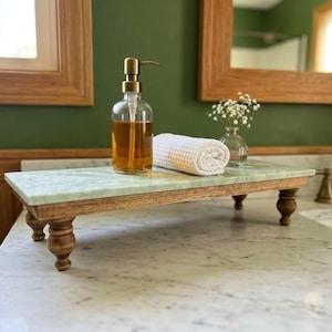May include: A rectangular riser featuring a light green marble top and a dark wood base with four turned legs. On the riser sits a clear glass soap dispenser with a gold pump filled with amber liquid and a white rolled waffle-weave towel.