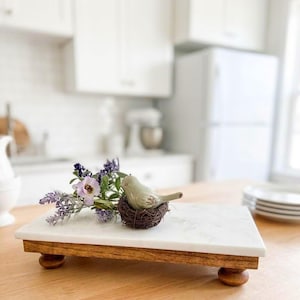 May include: A rectangular white marble serving board with a smooth surface, supported by four dark brown wooden ball feet. A small decorative light green ceramic bird sits in a brown twig nest next to a sprig of purple and white artificial flowers.
