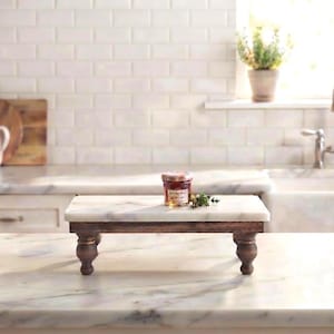 May include: A rectangular riser featuring a smooth white marble top surface supported by four dark brown turned wooden legs. A small glass jar with a label and a sprig of green herbs are placed on the center of the marble platform.