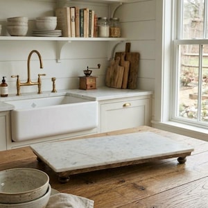 May include: A rectangular white marble serving board with grey veining, supported by four small dark wooden feet. The board is positioned on a wooden surface in a kitchen setting, featuring a smooth polished top surface and a sturdy base.