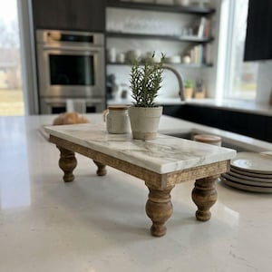 May include: A rectangular riser featuring a white marble top with grey veining, supported by four dark brown turned wooden legs. A small potted green plant and a small ceramic jar with a wooden lid sit on the marble surface.