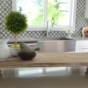May include: A rustic wooden tray with four legs, featuring a white pitcher, a small potted green plant, and a gray bird figurine in a nest. The tray is sitting on a gray countertop in a kitchen.
