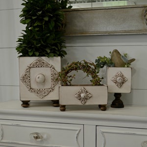 May include: Three white painted wooden boxes with decorative details and turned legs. The boxes are arranged on a white dresser with a drawer. The boxes are decorated with a floral design and a knob in the center. The boxes are perfect for storing small items or displaying decorative items.