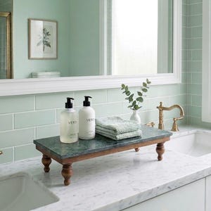 May include: A rectangular dark green marble riser with four turned wooden legs. On top sit two clear pump bottles with white labels reading VERD, and a stack of two folded light green textured towels next to a small white vase with green leaves.