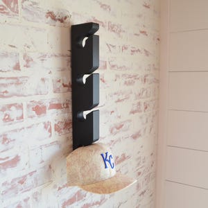 May include: Black wooden hat rack with four hooks mounted on a white brick wall. A beige baseball cap with a blue "KC" logo is hanging on the bottom hook.