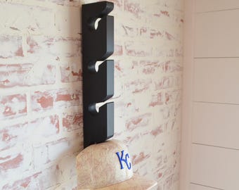 Puede incluir: Un perchero de sombreros de madera negro con cuatro ganchos montado en una pared de ladrillo blanco. Una gorra de béisbol beige con un logotipo "KC" azul está colgada en el gancho inferior.