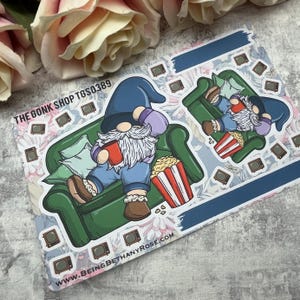 Puede incluir: Dos gnomos están relajándose en un sofá verde, viendo la televisión y disfrutando de palomitas de maíz. Los gnomos llevan pantalones vaqueros azules y un sombrero azul. El sofá tiene un cojín blanco y un cubo de palomitas de maíz a rayas rojas y blancas. El fondo es un patrón floral con el texto "THE GONK SHOP TGS0389" y "WWW.BEINGBETHANYROSE.COM".