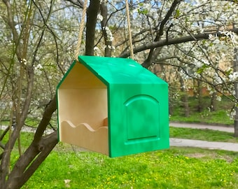 May include: A bright green birdhouse with a house-like shape, hanging from a tree branch. The birdhouse has a light wood interior and a decorative arched panel on the front. The bird feeder is suspended by a natural rope.