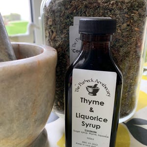 May include: A dark brown glass bottle with a black label that reads "The Purbeck Apothecary Thyme & Liquorice Syrup". The label also includes the ingredients "sugar syrup, thyme, liquorice", the contents "100ml", the expiry date "BBE 01.2022", and instructions to "Keep in cool dark place". The bottle is sitting on a yellow and white floral tablecloth.