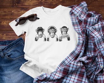 May include: White t-shirt featuring a graphic of three monkeys. The monkeys are wearing VR goggles, headphones, and holding a hamburger. The t-shirt is laid out on a wooden surface with a pair of sunglasses, a blue denim jacket, and a checked shirt.