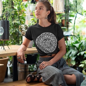 May include: A black t-shirt with a white graphic of the Aztec calendar. The person wearing the shirt is sitting on a wooden bench.