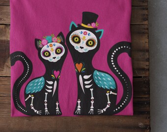 Könnte beinhalten: Ein pinkfarbenes T-Shirt mit einer schwarz-weißen Illustration von zwei Katzen, die in Day-of-the-Dead-Kleidung gekleidet sind. Die Katzen haben Zuckerköpfe und Blumenschmuck.