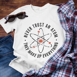 Puede incluir: Camiseta blanca con un gráfico negro y rojo de un átomo con el texto "Nunca confíes en un átomo. Ellos componen todo."