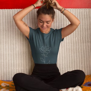 May include: A woman wearing a teal blue t-shirt with a white line drawing of a flower on the front. She is sitting on a white wicker sofa with a colourful blanket.