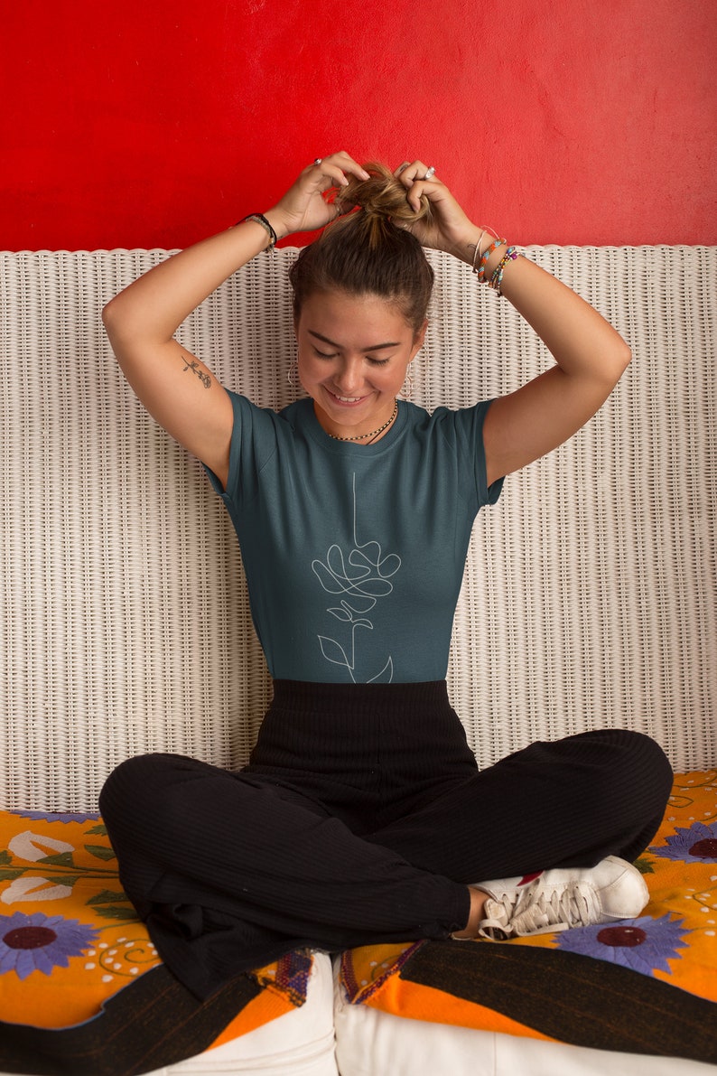 May include: A woman wearing a teal blue t-shirt with a white line drawing of a flower on the front. She is sitting on a white wicker sofa with a colourful blanket.