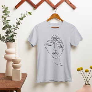 Puede incluir: Una camiseta azul claro con un dibujo de línea negra de una cara con una rama frondosa que crece de la frente.