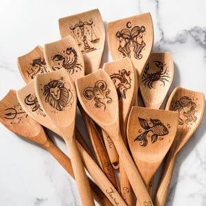 Pode incluir: Um conjunto de 12 colheres de madeira com signos do zodíaco gravados na frente de cada colher. As colheres estão dispostas em uma pilha com as alças para fora. Os signos do zodíaco são: Áries, Touro, Gêmeos, Câncer, Leão, Virgem, Libra, Escorpião, Sagitário, Capricórnio, Aquário e Peixes.
