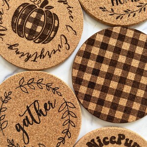 May include: A selection of circular cork coasters with autumnal designs. One coaster displays a pumpkin with a plaid pattern, while another says "gather" in script. Other coasters have the phrases "spice & everything" and "nice pumpkin".