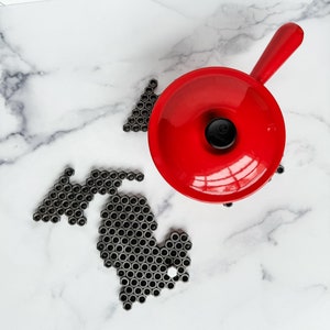 Op de afbeelding: Een felrode steelpan met een lange handgreep staat op een marmeren oppervlak. Naast de pan staat een gedetailleerde omtrek van de staat Michigan, gemaakt van talrijke kleine, donkergrijze metalen stukjes. De achtergrond is een witte tegel backsplash.